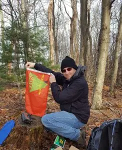 Eric holds up his SOTA flag while kneeling over his radio setup in the woods.